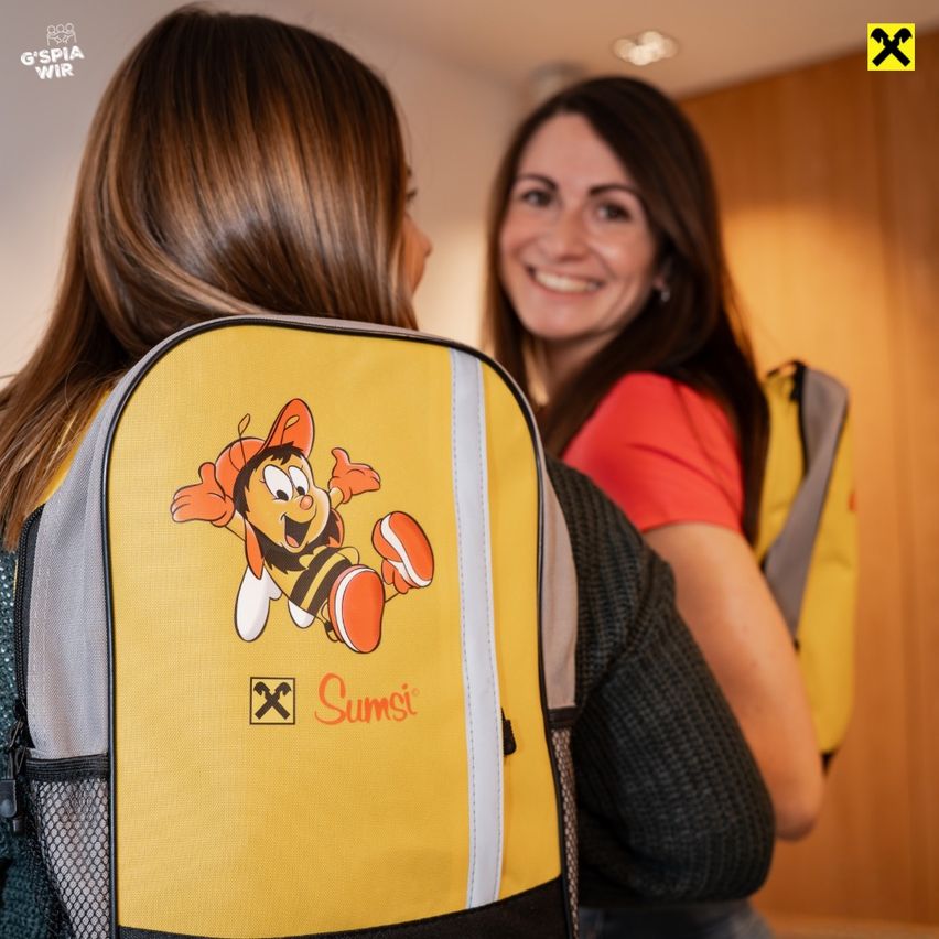 Zwei Frauen stehen neben einem gelben Rucksack mit einem Cartoon-Bienencharakter darauf. Die Frau auf der linken Seite schaut auf den Rucksack, während die Frau auf der rechten Seite lächelt.