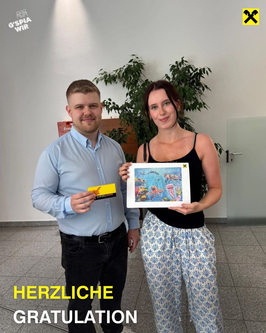 Ein Mann und eine Frau stehen in einem Raum, lächeln. Der Mann hält eine gelbe Karte und die Frau hält ein Bild mit einer Ozeanszene. Der Raum hat weiße Wände, eine Pflanze und eine Tür.