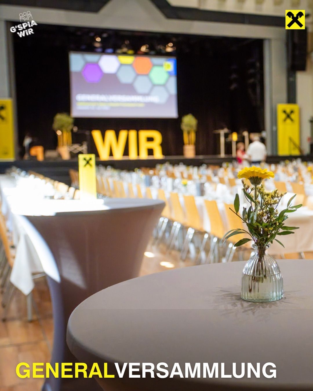 Ein Konferenzsaal ist eingerichtet mit Tischen und Stühlen. Eine Leinwand zeigt eine Präsentation. Ein Tisch hat eine Vase mit Blumen. Das Wort 'WIR' ist prominent auf der Bühne zu sehen.