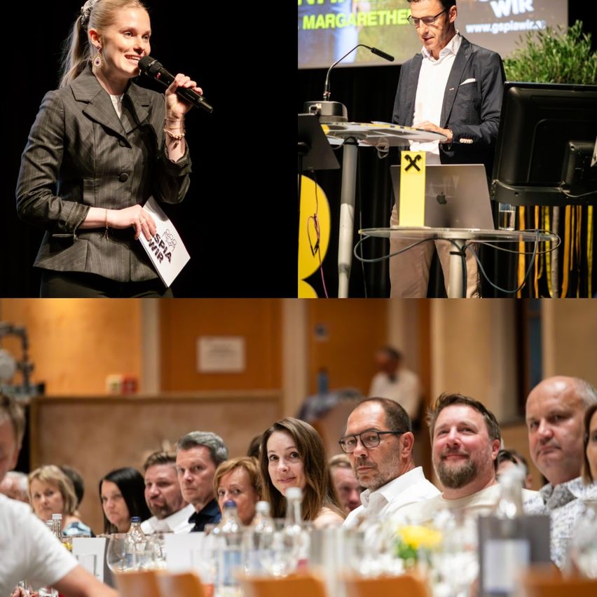 Eine Frau hält ein Mikrofon und Papier in der Hand, während sie auf der Bühne steht. Ein Mann steht an einem Podium mit Laptop und Mikrofon. Unten sitzt eine Gruppe von Menschen an einem Tisch mit Weinflaschen.