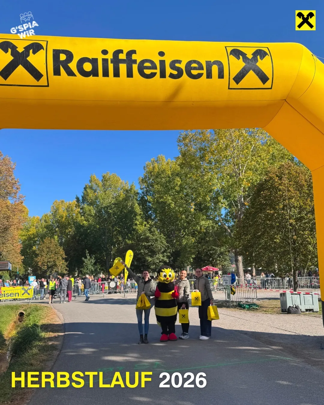 Vier Personen stehen unter einem gelben Bogen mit dem Text Raiffeisen und Herbstlauf 2026. Sie sind in Kostümen gekleidet und halten gelbe Taschen.
