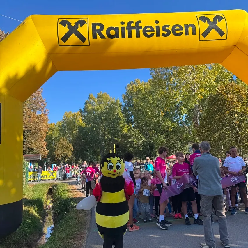 Ein Maskottchen, das als Biene verkleidet ist, steht vor einem gelben Bogen mit dem Wort Raiffeisen. Hinter dem Maskottchen stehen mehrere Menschen und beobachten einen Marathon.