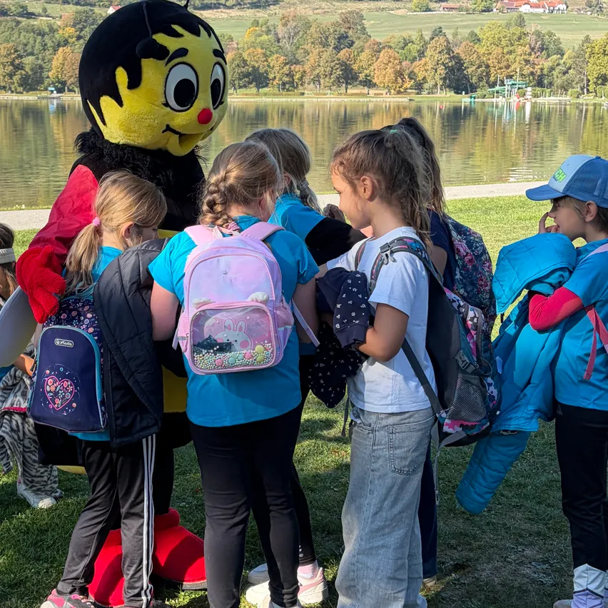 Eine Gruppe junger Kinder versammelt sich um eine große Biene-Mascotte auf einem Grasfeld in der Nähe eines Sees.