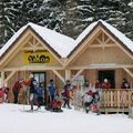Skifahrer und Snowboarder versammeln sich vor einer hölzernen Hütte namens Langlaufzentrum Jakob, mit schneebedeckten Dächern, verschneiten Tannen und Skiausrüstung auf der Veranda.