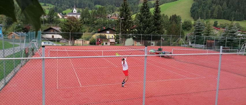 Ein Tennisspieler in einem roten Shirt und Shorts serviert einen Tennisball auf einem roten Tennisplatz mit Zaun. Im Hintergrund befinden sich Berge und Häuser.
