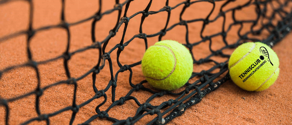 Zwei Tennisbälle liegen an einem schwarzen Netz auf einem Sandplatz.