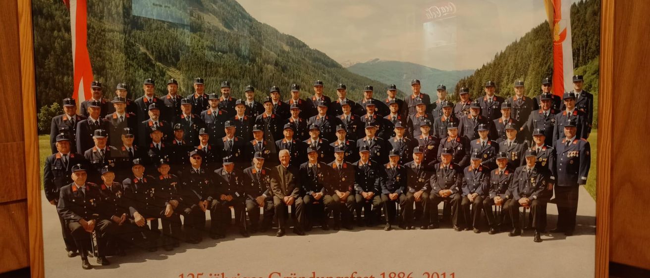Eine Gruppe von uniformierten Personen steht vor Bergen. Der Text lautet '125-jähriges Gründungsfest 1886-2011.'