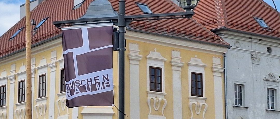 Eine Straßenlampe mit einer Fahne vor einem gelben Gebäude mit roten Dächern. Der Laternenpfahl hat einen Fahnenmast mit bunten Bändern.