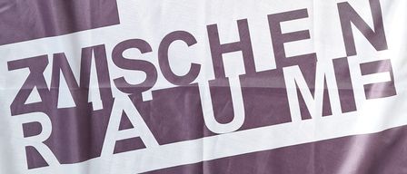 Ein lila und weißes Banner mit den Worten 'Zwischen Räume' in Weiß, auf einer Holzoberfläche platziert.