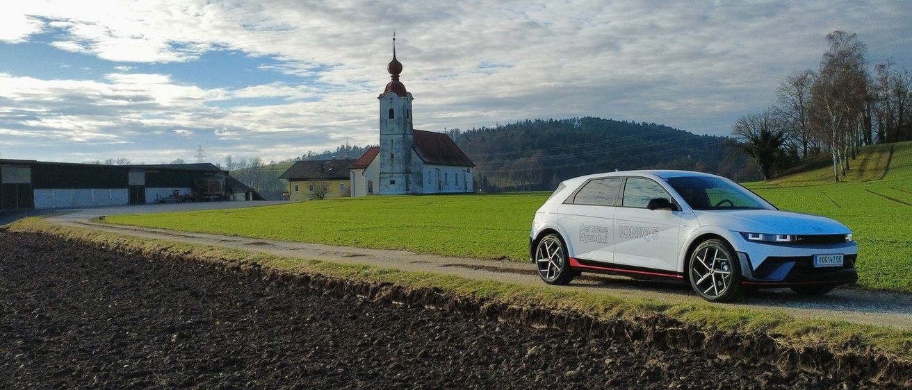 Ein weißes Auto fährt auf einer Landstraße. Im Hintergrund befindet sich eine Kirche mit einem Turm und einem kleinen Haus. Der Himmel ist blau mit Wolken und es gibt ein grasbewachsenes Feld.