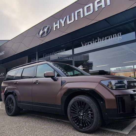 Ein brauner Hyundai SUV steht vor einem Hyundai-Händler mit der Aufschrift 'Versicherung' auf der Scheibe.