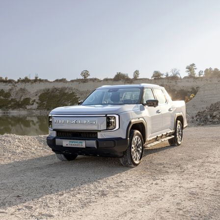 Ein weißer elektrischer Pickup-Truck, der Maxus eTeron, steht auf einer staubigen Straße in der Nähe eines Gewässers.