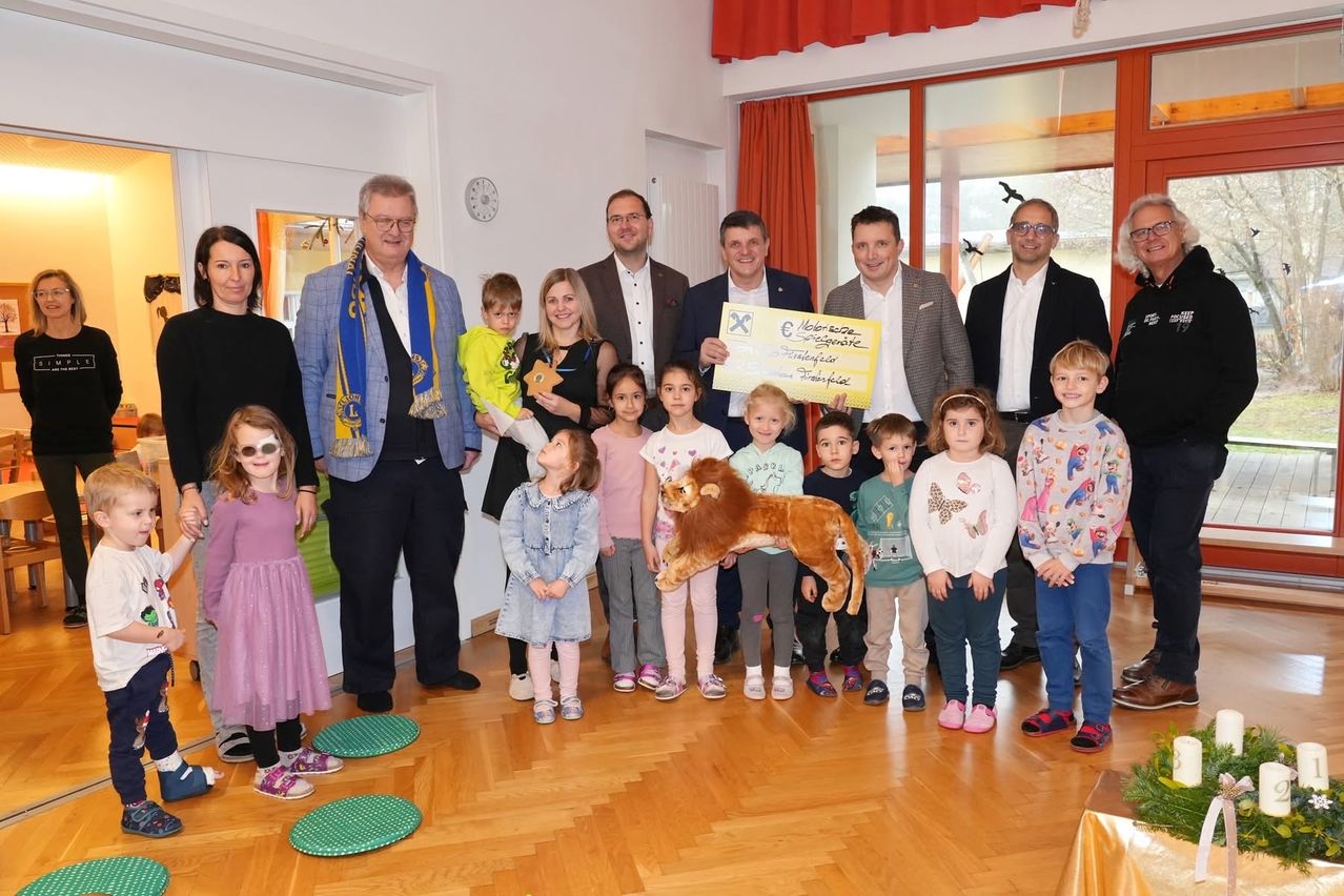 Eine Gruppe von Erwachsenen und Kindern posiert für ein Foto in einem Raum, wobei ein Mann einen großen Scheck und einen pelzigen Löwen hält.