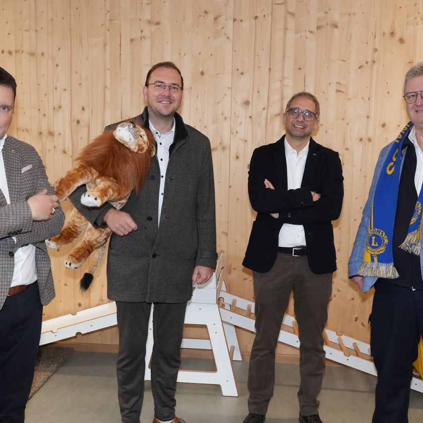 Vier Männer stehen lächelnd vor einer Holzwand. Einer hält ein Löwenplüschtier. Ein anderer verschränkt die Arme, während ein Dritter einen Schal trägt. Der Vierte hat einen Gürtel. Hinter ihnen stehen weiße Klappstühle.