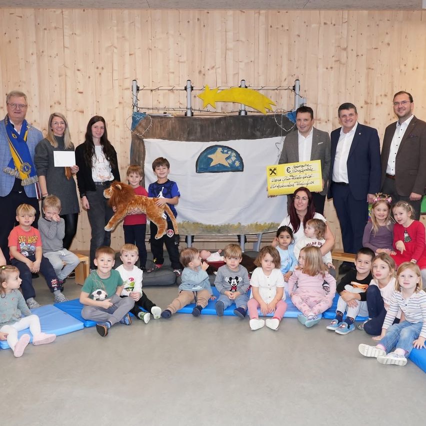 Eine Gruppe von Erwachsenen und Kindern posiert für ein Foto in einem Raum mit Holzwänden, einige sitzen auf Matten, einer hält ein Kuscheltier, und ein Banner mit einem Stern dahinter.