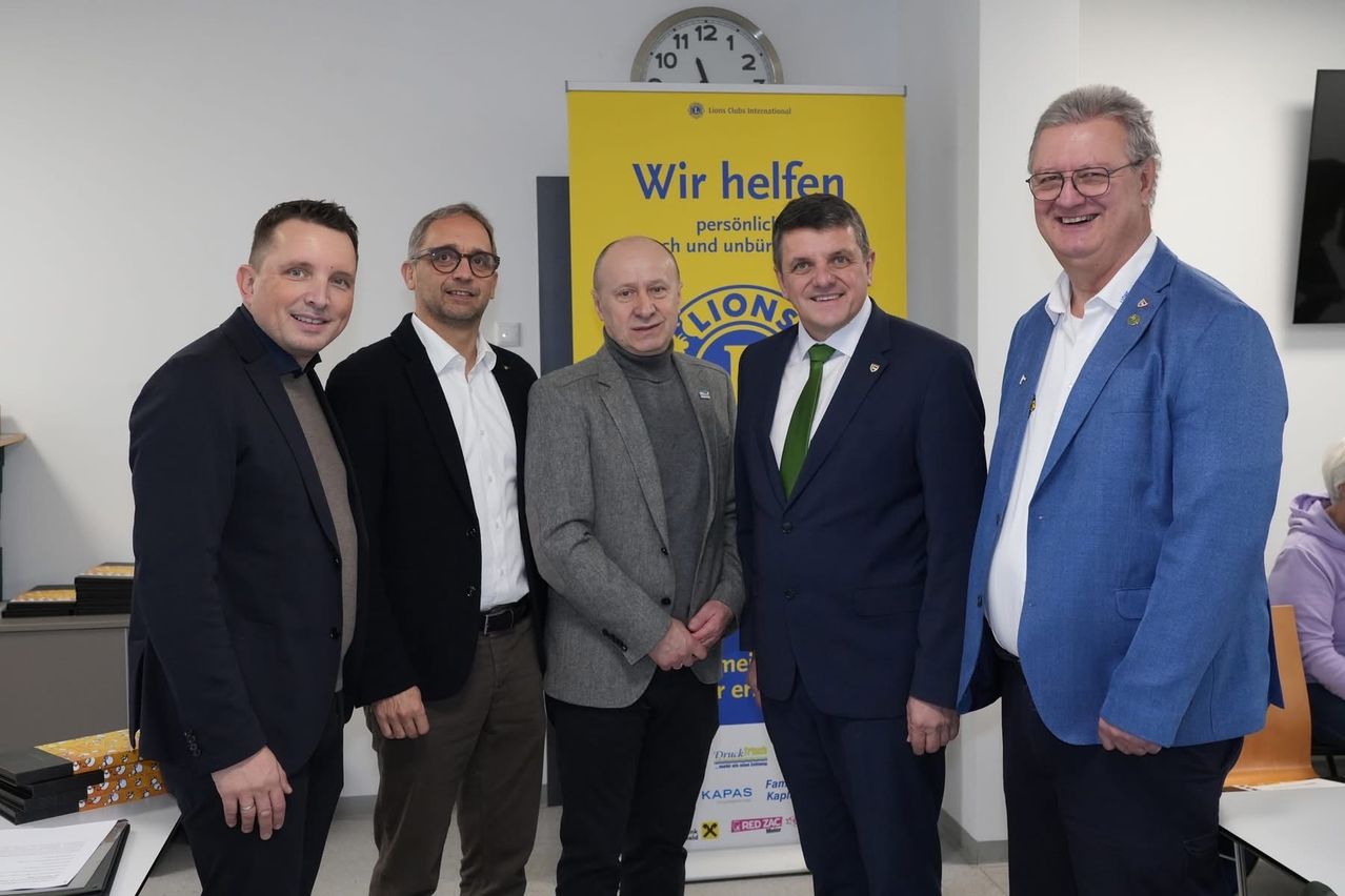 Fünf Männer stehen vor einem gelben Lions Club-Banner und lächeln. Der Mann ganz rechts trägt einen blauen Anzug und eine Brille.
