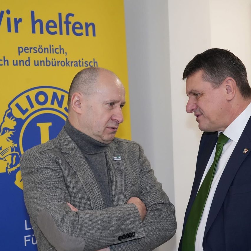 Zwei Männer stehen vor einem gelben Banner mit den Worten 'Wir helfen' in blauen Buchstaben. Der Mann auf der linken Seite trägt einen grauen Mantel, während der Mann auf der rechten Seite einen blauen Anzug mit einer grünen Krawatte trägt.