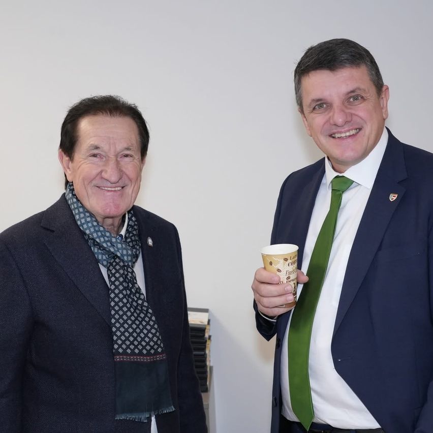 Zwei Männer stehen in einer Innenumgebung zusammen und lächeln. Der Mann auf der linken Seite trägt einen Schal und eine Abzeichen. Der Mann auf der rechten Seite hält einen Kaffeebecher und trägt eine grüne Krawatte.