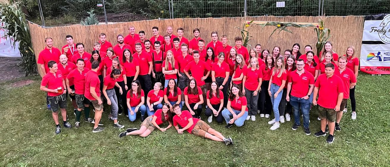 Eine Gruppe von Menschen in roten Shirts und verschiedenen Kleidungsstücken posiert für ein Foto auf einer Rasenfläche mit einem Holzzaun und Bäumen im Hintergrund.