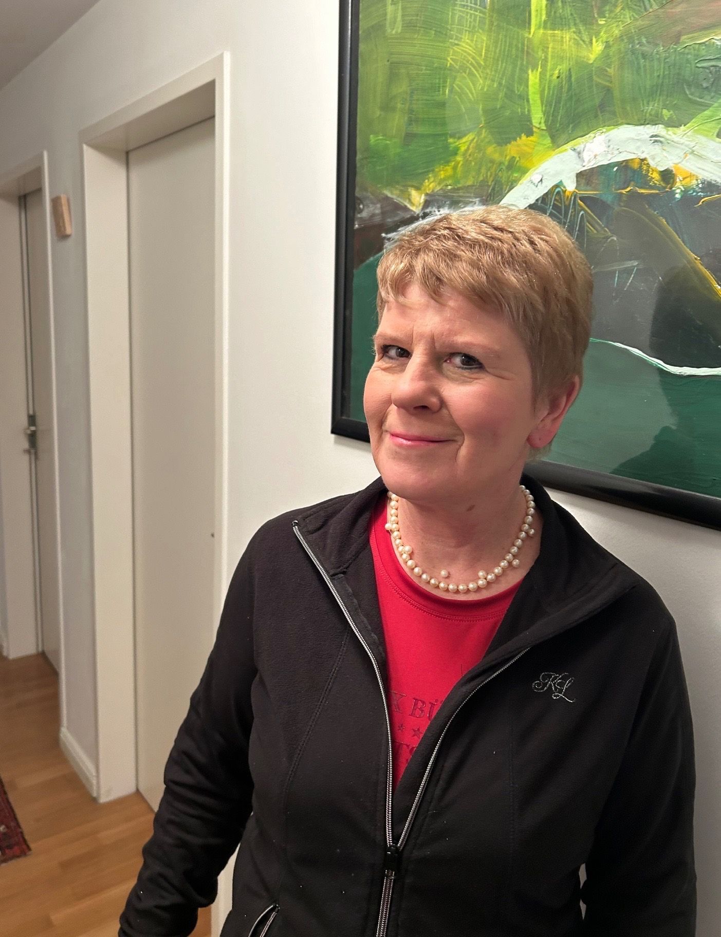 Eine Frau in einem schwarzen Jackett und rotem Shirt mit einer Perlenkette steht vor einem farbenfrohen abstrakten Gemälde. Hinter ihr befinden sich zwei weiße Türen.