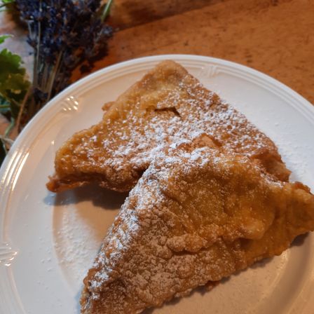 Zwei Stücke frittierter Teig mit Puderzucker bestreut liegen auf einem weißen Teller mit einem braunen Holzhintergrund und einem Lavendelzweig.