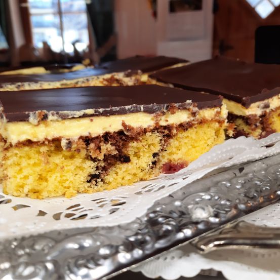 Ein Stück Marmorkuchen mit Schokoladenüberzug liegt auf einem silbernen Teller, der auf einer Spitze platziert ist. Der Kuchen hat eine weiße und braune Schicht mit einem Hauch von Rot.
