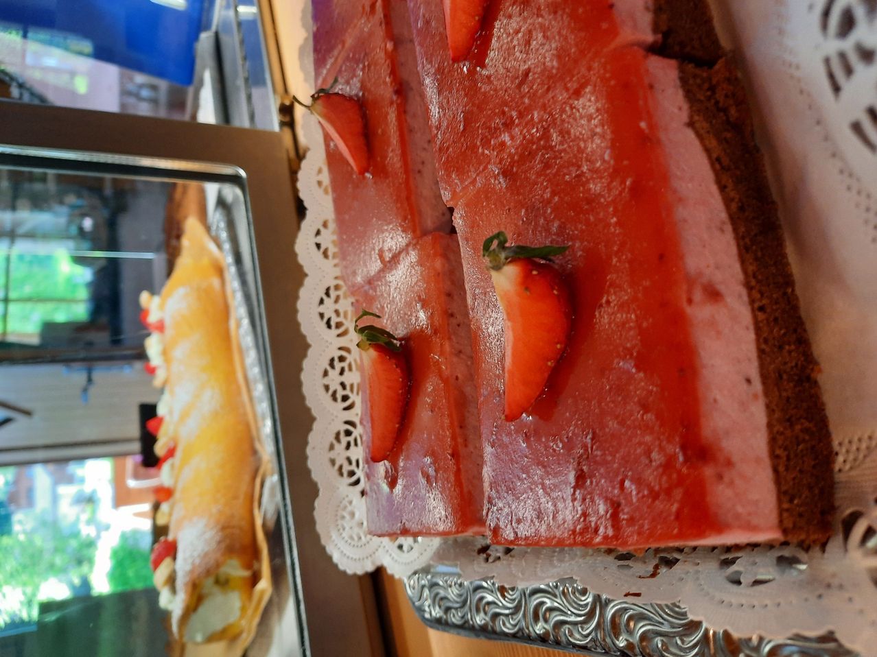 Ein Dessert mit roter Gelee-Oberfläche und Erdbeeren auf einem weißen Spitzentablett. Im Hintergrund ist ein weiteres Dessert mit gelber Cremefüllung zu sehen.