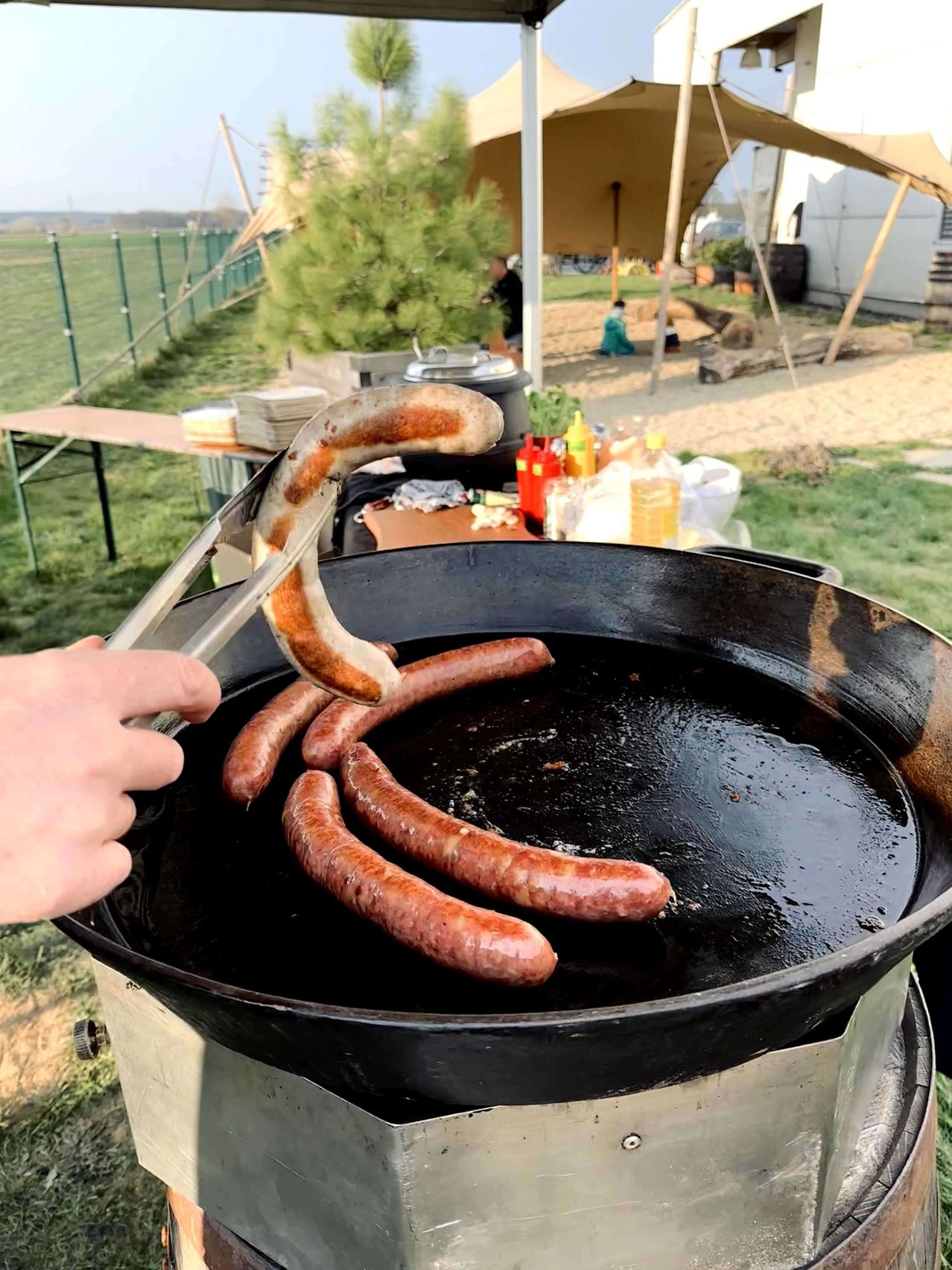 Eine Person benutzt eine Zange, um eine Wurst aus einer schwarzen Pfanne zu heben. Vier Würste werden in der Pfanne gekocht. Würzmittel stehen auf einem Tisch im Hintergrund.