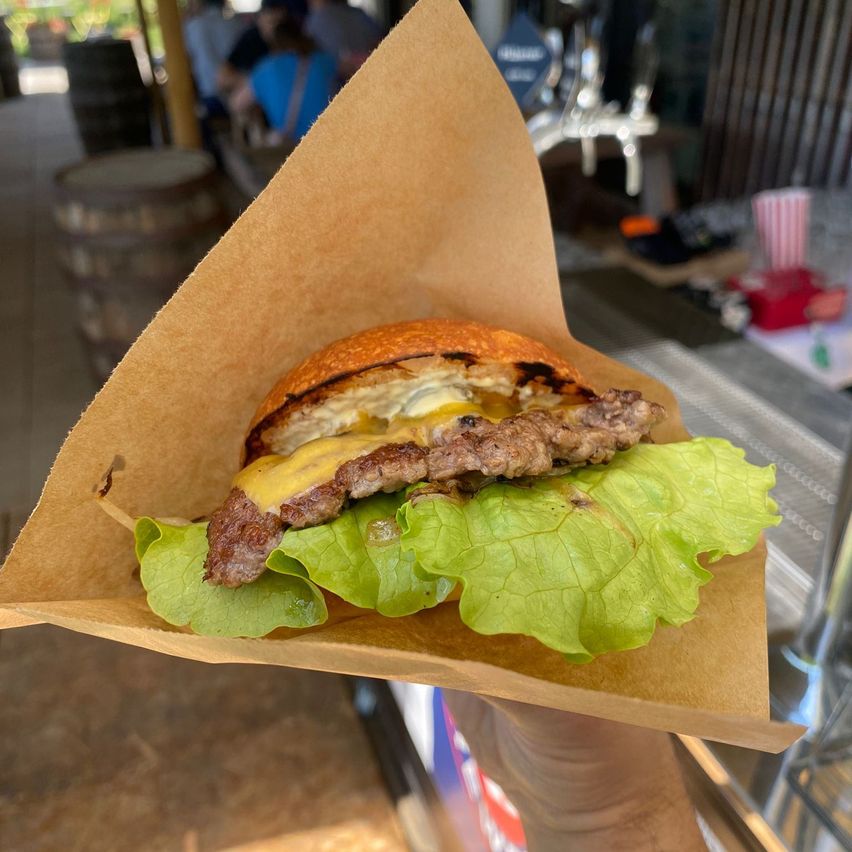 Eine Person hält einen Burger mit geschmolzenem Käse, Salat und einem gebratenen Ei, alles in einem braunen Papierkegel eingewickelt. Dahinter sind ein Fass, eine Theke und andere Gegenstände zu sehen.
