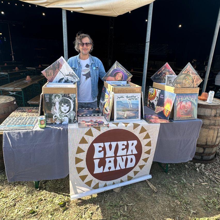 Ein Mann steht hinter einem Tisch, auf dem ein Banner mit der Aufschrift Ever Land befestigt ist. Der Tisch ist mit verschiedenen Schallplatten und Kisten sowie einigen Dosen bestückt. Hinter ihm sind Tische und Bänke unter einem Zelt aufgestellt.