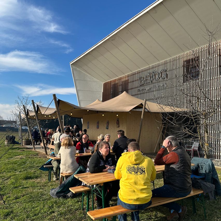 Eine Gruppe von Menschen sitzt unter einem Vordach um Tische herum in einem Außenbereich, wahrscheinlich in einem Restaurant, mit einem modernen Gebäude im Hintergrund.