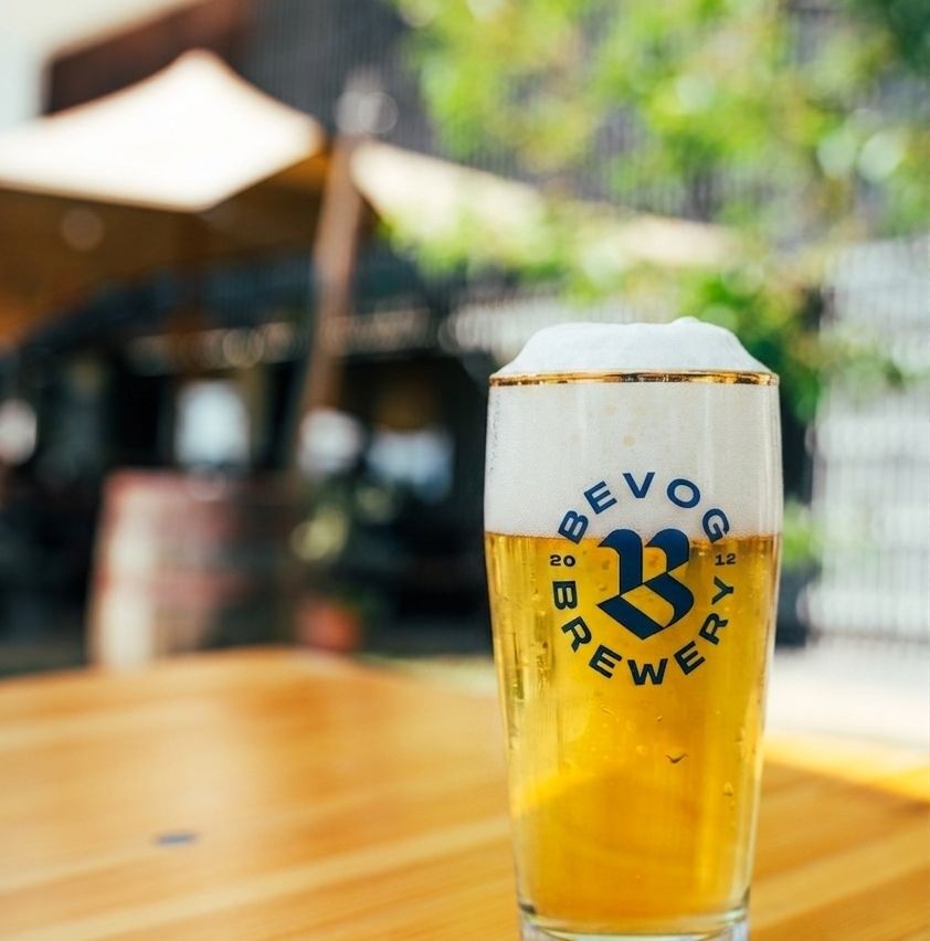 A glass of beer with a frothy head, labeled 'BE VO 20 BREWERY 12', on a wooden table with a blurry background of a bar.