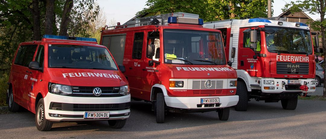 Drei Feuerwehrfahrzeuge sind auf einem Parkplatz geparkt. Zwei der Fahrzeuge sind rot und weiß und eines ist weiß. Das Wort 'FEUERWEHR' steht auf der Front der roten Fahrzeuge.