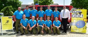 Eine Gruppe junger Männer in blauen Uniformen posiert für ein Foto vor einem roten Fahrzeug mit einem Mann in weißem Hemd und Krawatte.