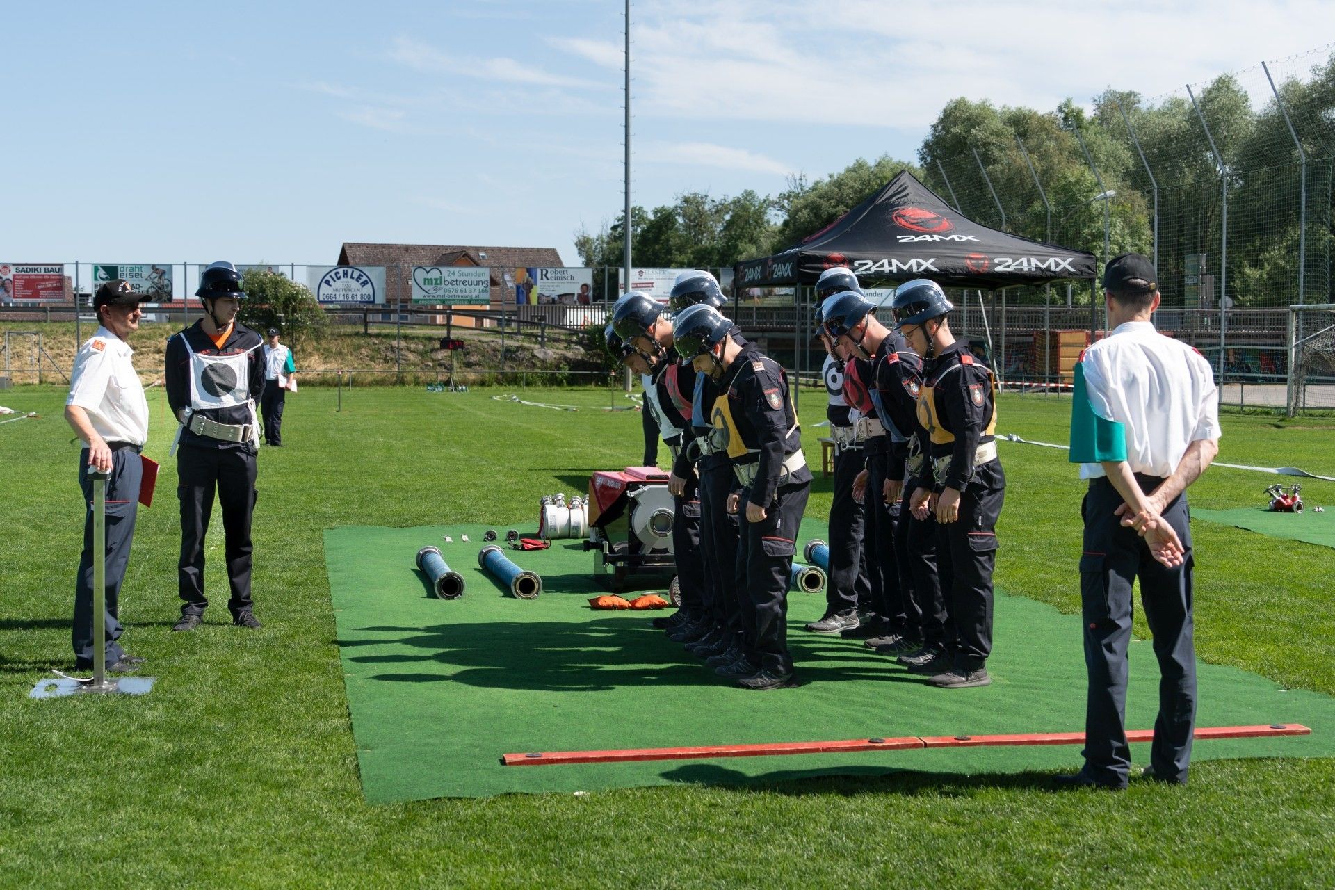 Eine Gruppe von Menschen in Helmen und Uniformen steht auf einem grünen Feld, möglicherweise in Vorbereitung auf eine Sportveranstaltung. Ausrüstung und ein Zelt sind in der Nähe.