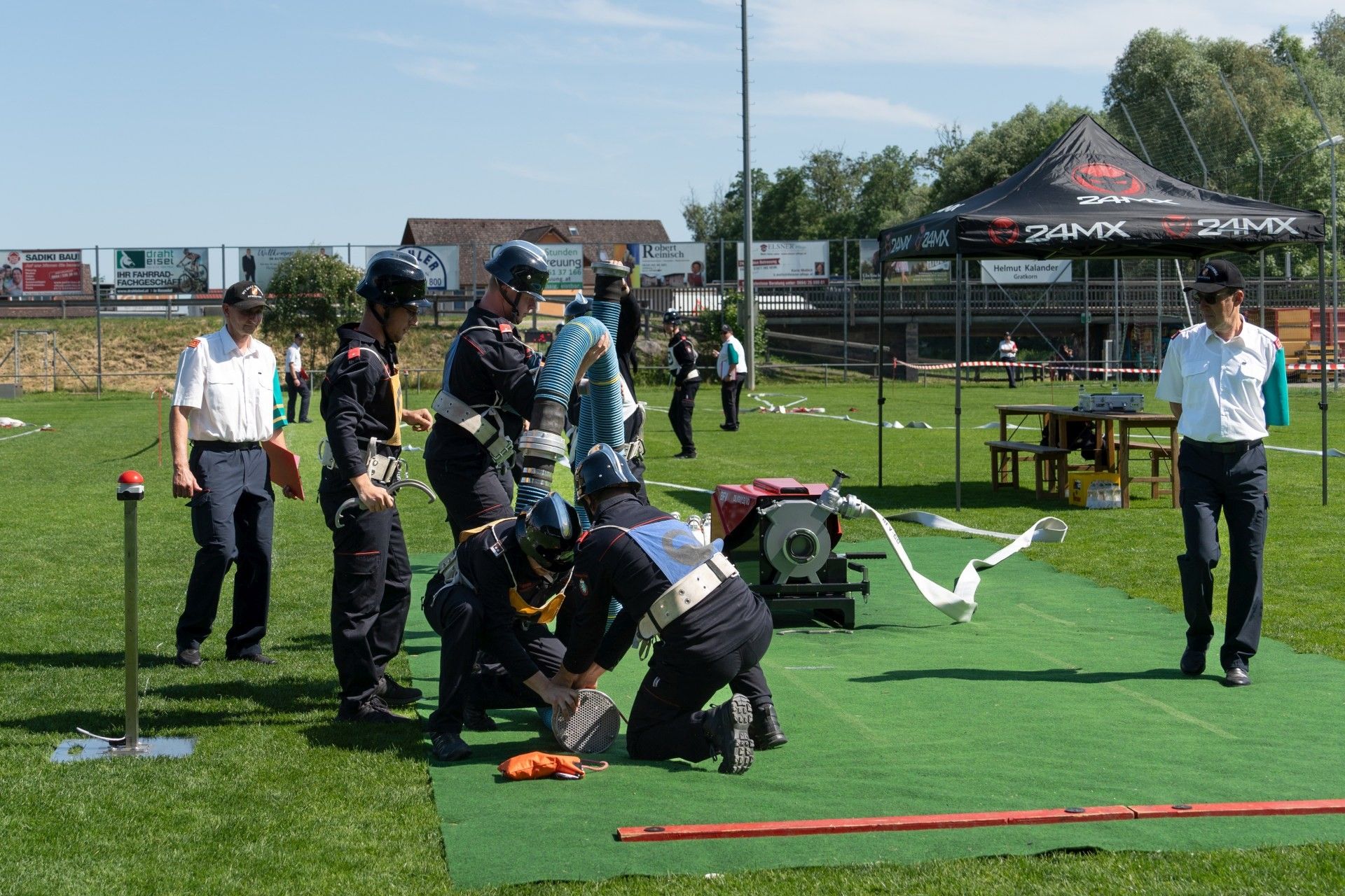 Rettungskräfte üben auf einem Feld. Sie haben Ausrüstung und eine Maschine in der Nähe. Ein Zelt und Tische sind mit Gegenständen aufgebaut. Im Hintergrund sind Menschen und ein Gebäude zu sehen.