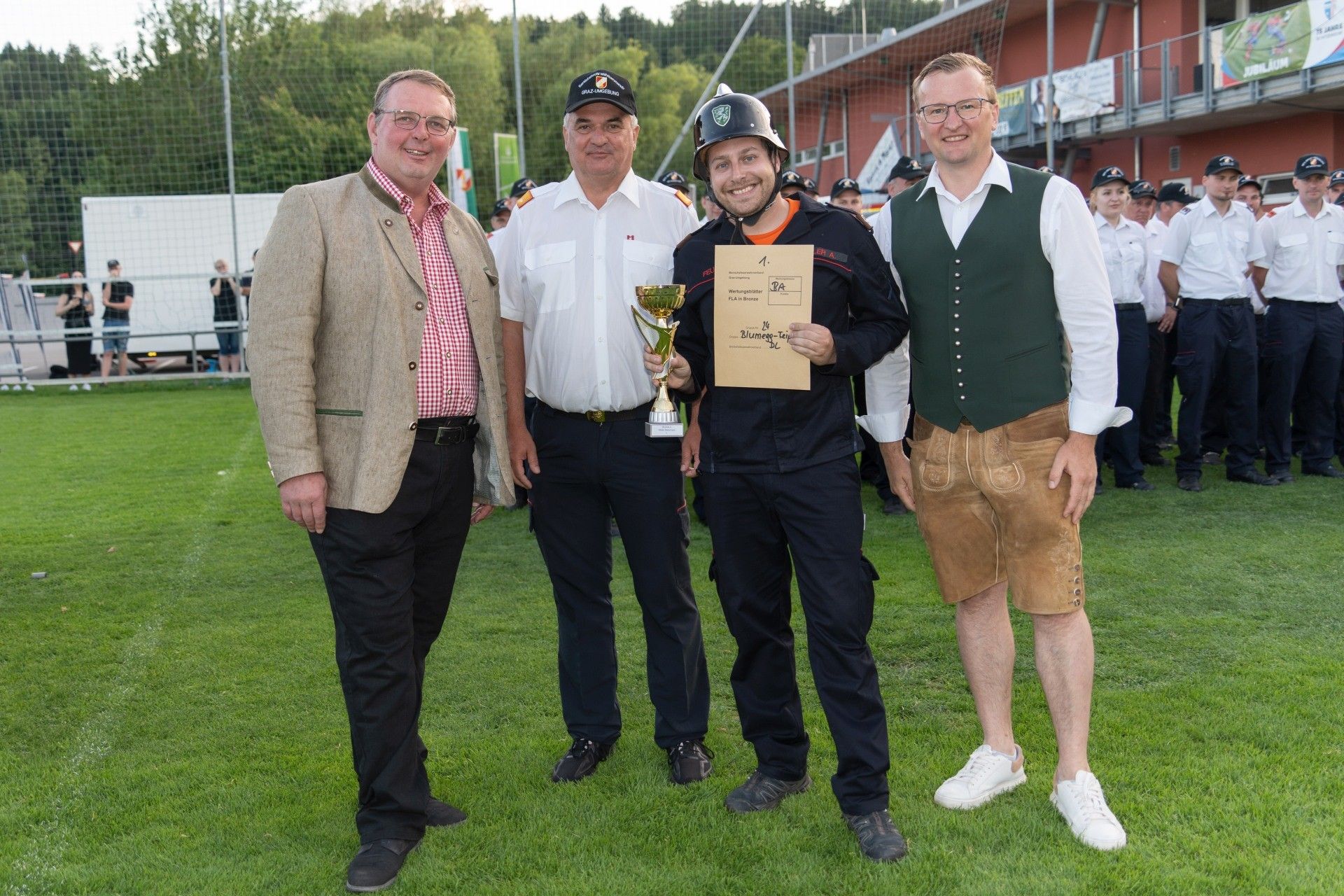 Vier Männer stehen auf einem grünen Feld, zwei vorne mit Brille und lächelnd, einer in einem Hut mit Trophäe und Urkunde und einer in traditioneller Kleidung.