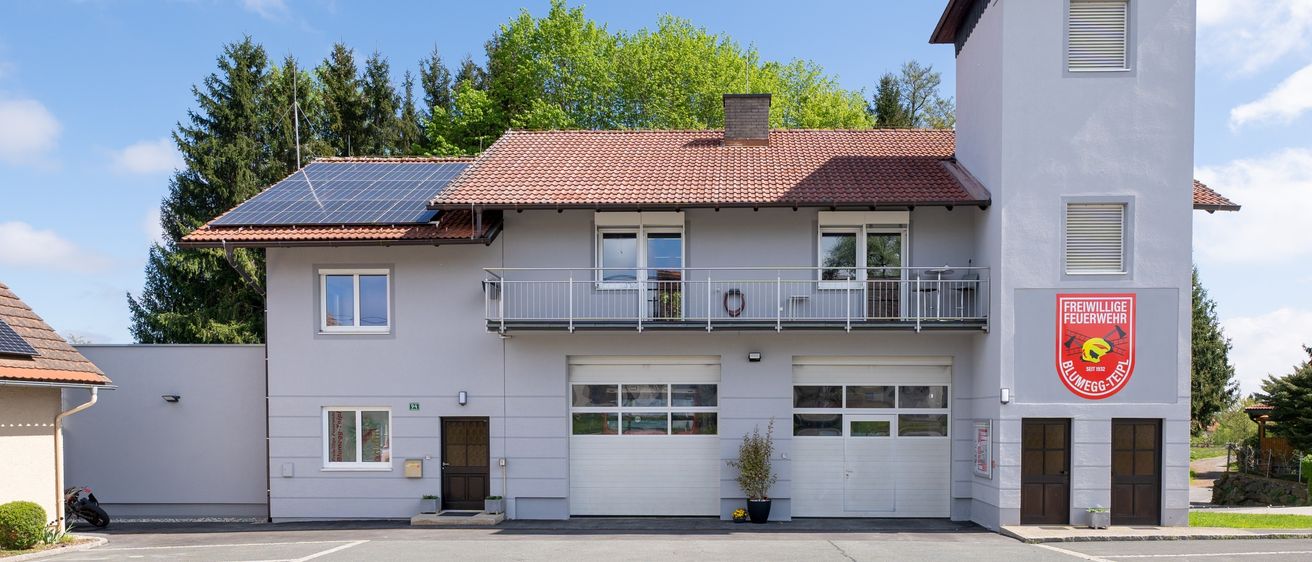 Ein modernes zweistöckiges Haus mit rotem Dach und Solarpanelen. Es gibt zwei Garagen, von denen eine eine Terrasse mit Geländer hat. Ein Feuerwehremblem ist an der Wand angebracht.