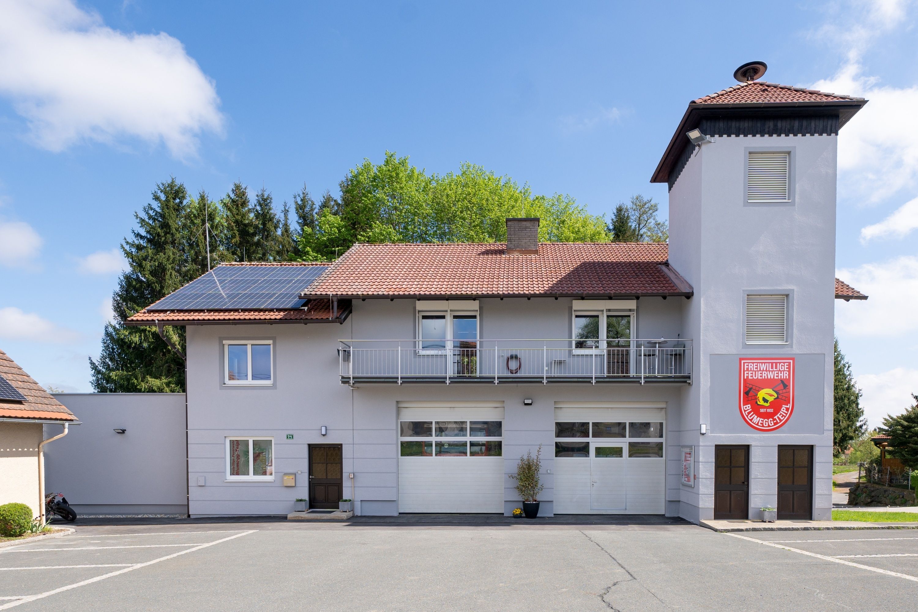 Ein Feuerwehrhaus mit rotem Dach und Solarmodulen. Es gibt zwei Garagen und einen Balkon mit Geländer. Die Vorderseite hat ein rotes Schild mit weißem Text.