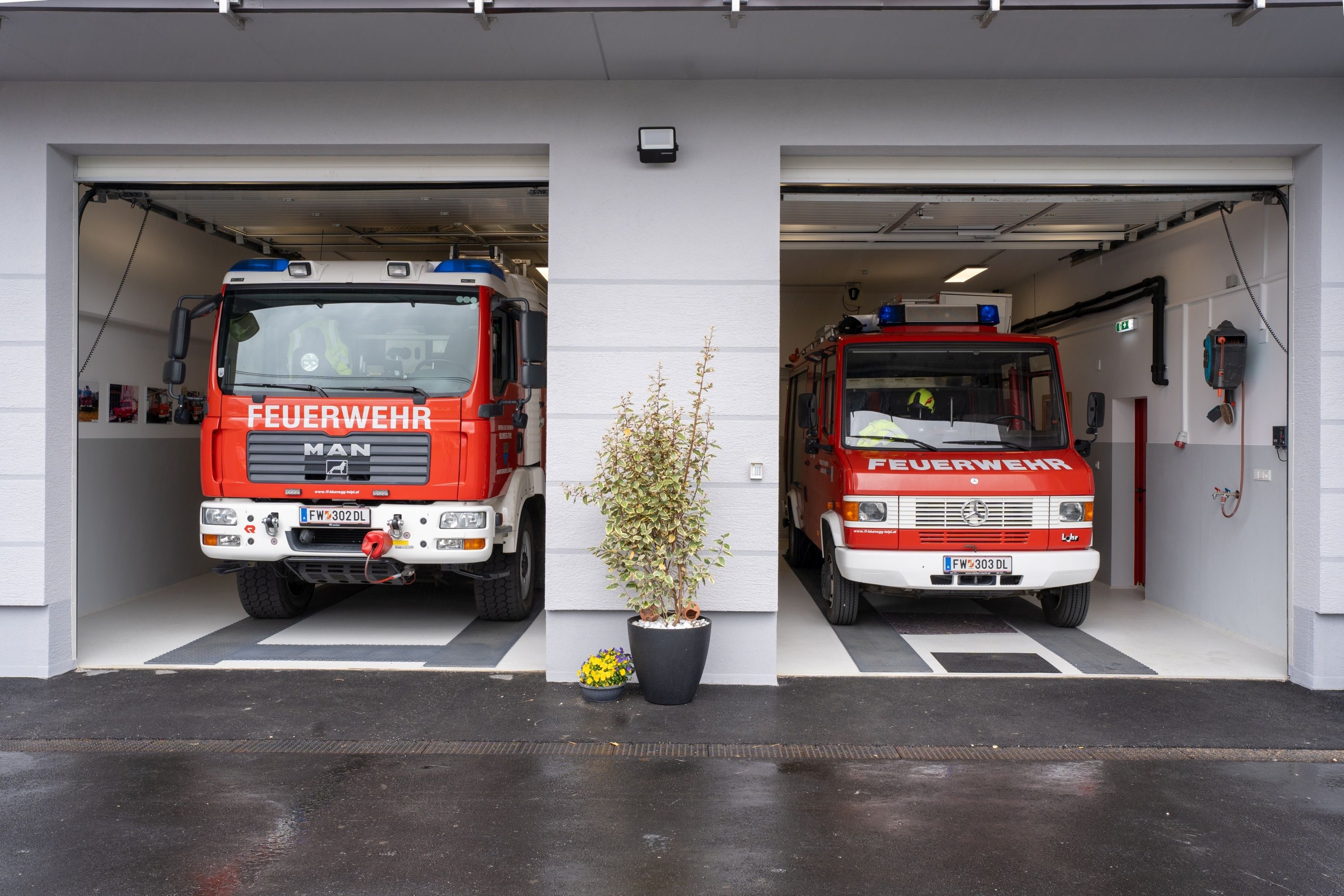 Zwei rote Feuerwehrfahrzeuge sind in einer Garage mit einer Pflanze und gelben Blumen geparkt. Die Fahrzeuge haben blaue Lichter auf dem Dach und 'FEUERWEHR' steht an der Seite.