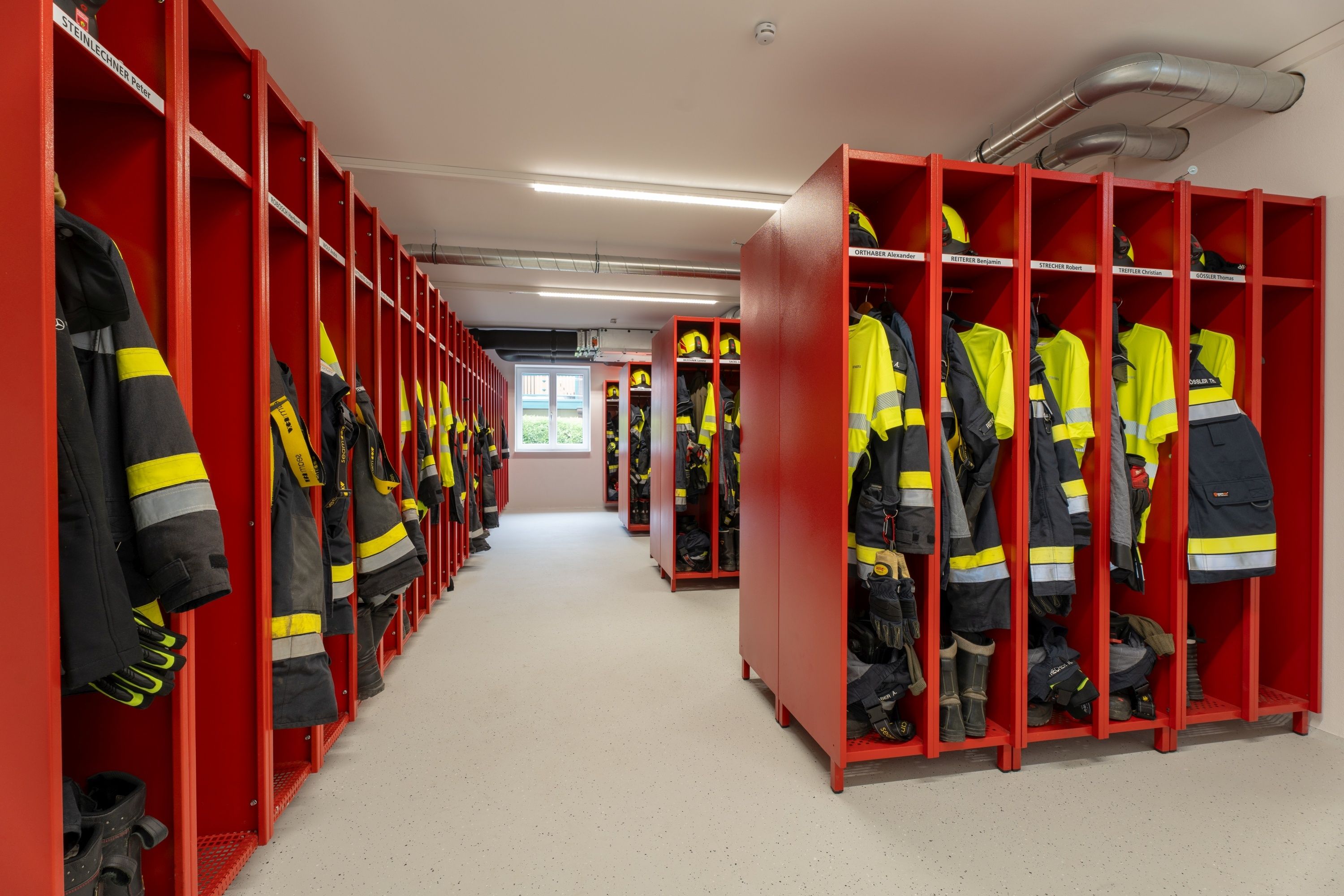 Ein Flur mit roten Spinden an beiden Seiten, gefüllt mit gelben und schwarzen Feuerwehruniformen. Rechts befinden sich zwei Reihen von Spinden mit mehreren Uniformen. Links ist eine einzelne Reihe von Spinden zu sehen. Die Decke hat Lampen und eine Rohrleitung.