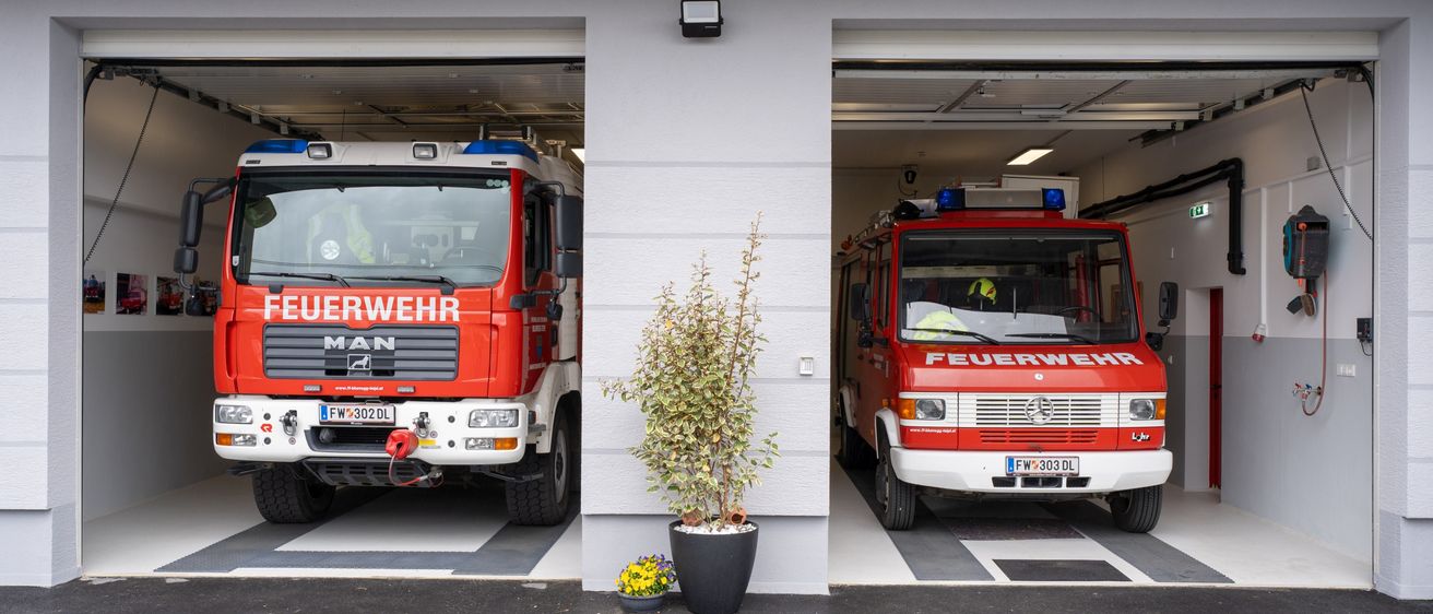 Zwei rote Feuerwehrfahrzeuge sind in einer Feuerwache geparkt. Ein Topf steht zwischen den Fahrzeugen. Die Fahrzeuge haben das Wort 'Feuerwehr' darauf geschrieben.