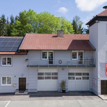Ein modernes Haus mit rotem Dach und Solarpanelen. Es hat zwei Garagen und einen Balkon mit Geländer. Im Hintergrund befinden sich Bäume.