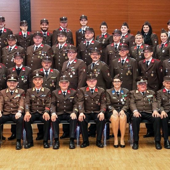 Eine Gruppe uniformierter Personen, wahrscheinlich Militär oder Polizei, ist für ein Foto versammelt. Sie sitzen in Reihen auf einem Holzboden, einige stehen. Sie tragen Hüte, Krawatten und Medaillen.