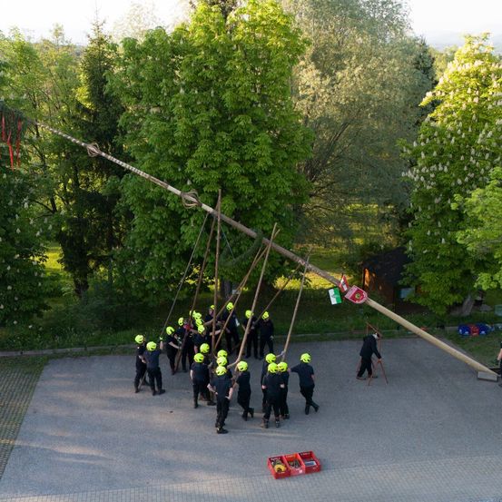 Eine Gruppe von Menschen mit gelben Helmen hebt einen langen Pfahl in einem Außenbereich. Der Pfahl ist mit Seilen gefesselt. Der Bereich ist von Bäumen umgeben. Es gibt ein Gebäude mit einer Flagge an der Seite. Es gibt eine rote Aufbewahrungsbox auf dem Boden.