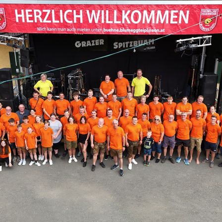 Eine Gruppe von Menschen in orangefarbenen Shirts steht vor einer Bühne mit einem roten Banner, das Herzlich Willkommen steht. Dahinter stehen zwei Personen auf der Bühne mit einem Mikrofon.