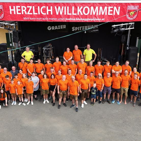 Eine Gruppe von Menschen in orangefarbenen Shirts steht vor einer Bühne mit einem roten Banner, das Herzlich Willkommen steht. Dahinter stehen zwei Personen auf der Bühne mit einem Mikrofon.