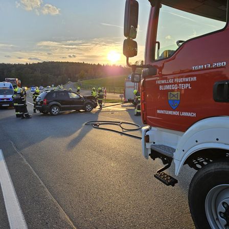 Rettungsfahrzeuge, einschließlich eines roten Feuerwehrwagens, sind an der Unfallstelle. Feuerwehrleute und Beamte sind anwesend, mit der untergehenden Sonne im Hintergrund.