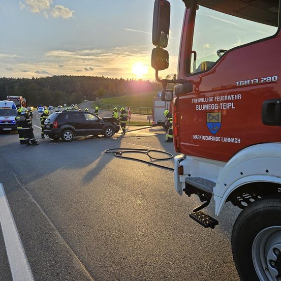 Rettungsfahrzeuge, einschließlich eines roten Feuerwehrwagens, sind an der Unfallstelle. Feuerwehrleute und Beamte sind anwesend, mit der untergehenden Sonne im Hintergrund.