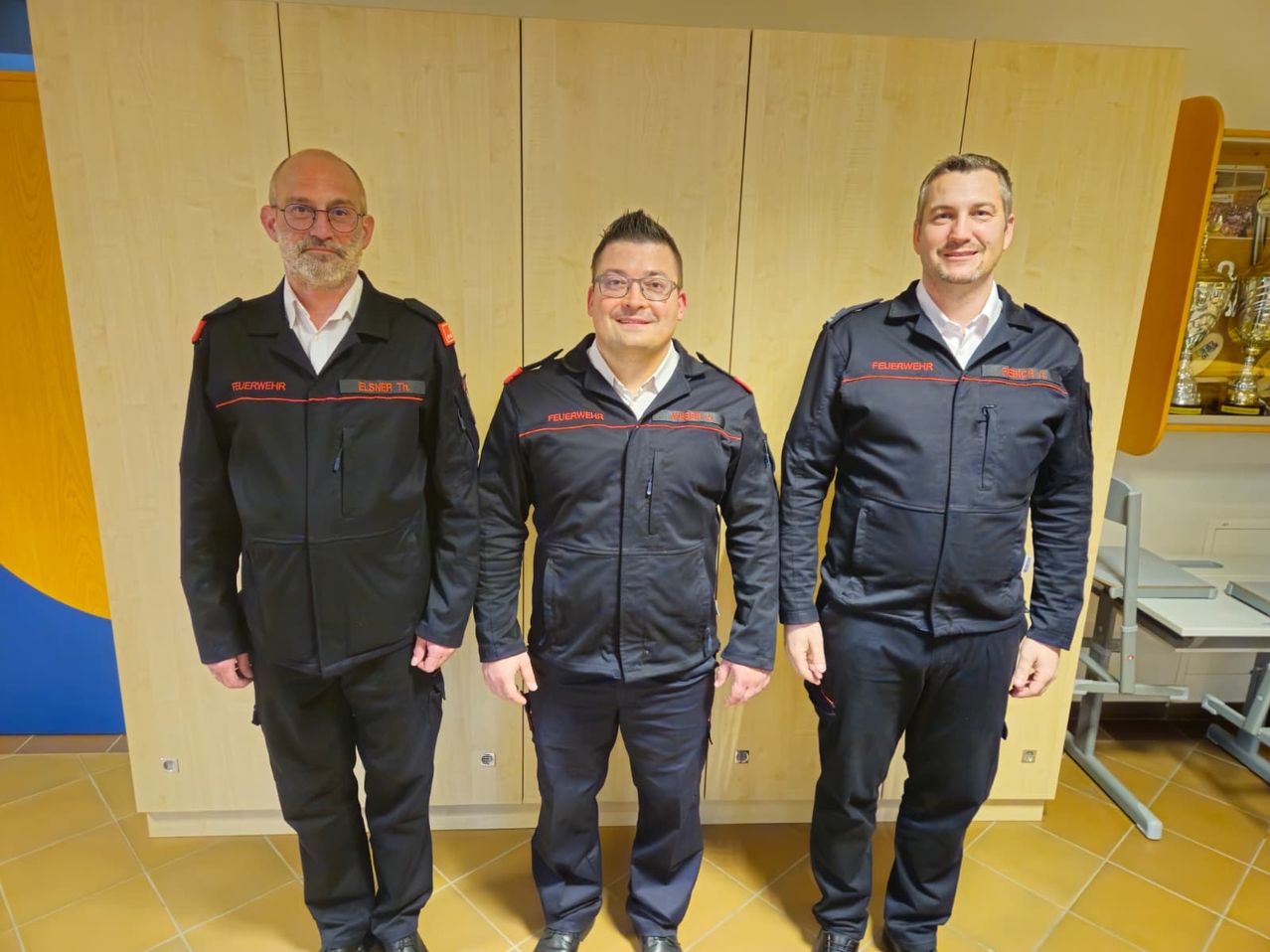 Drei Männer in schwarzen Uniformen stehen zusammen vor einer Holzwand und lächeln für ein Foto. Sie sind wahrscheinlich Feuerwehrleute, wie ihre Uniformen zeigen.